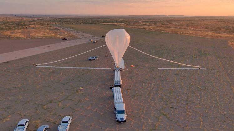 NASA Sounding Rocket Aims for Aug. 25 Launch 2 Aerial view of a high-altitude scientific balloon launch preparation in a desert setting at sunset. The white balloon is partially inflated and tethered to a launch vehicle, with support vehicles and personnel positioned around the launch area on a cracked desert surface