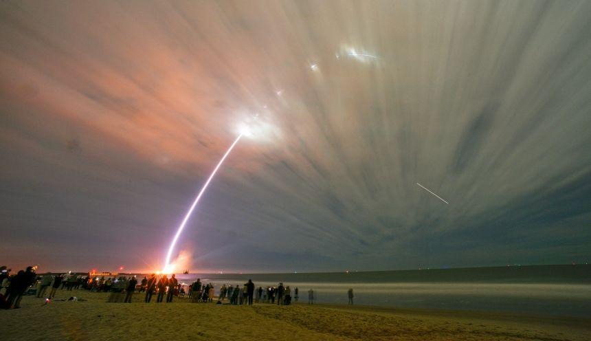 Spectacular launch of a rocket into the sky, captured from a beach as onlookers watch the ascent against a backdrop of clouds and vibrant colors.
