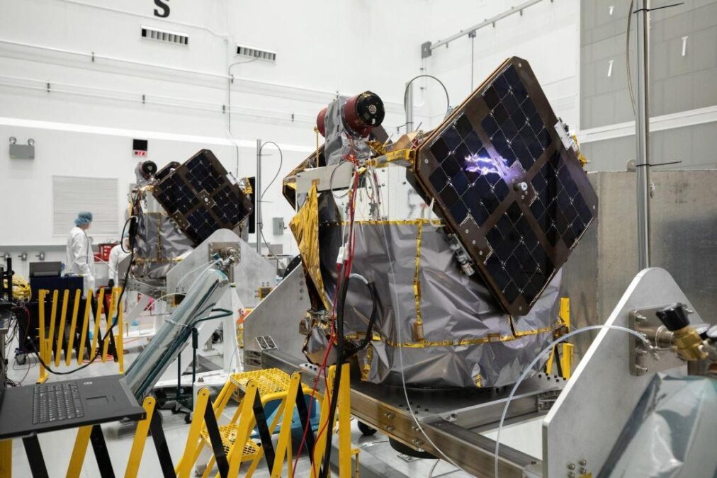 Two satellites with solar panels are shown in a cleanroom environment, being prepared for launch. A person is visible working in the background, surrounded by equipment and safety barriers.