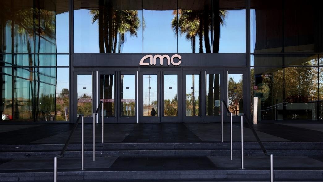 Entrance of an AMC theater showcasing the company’s logo on the glass doors with reflections of palm trees in the background.