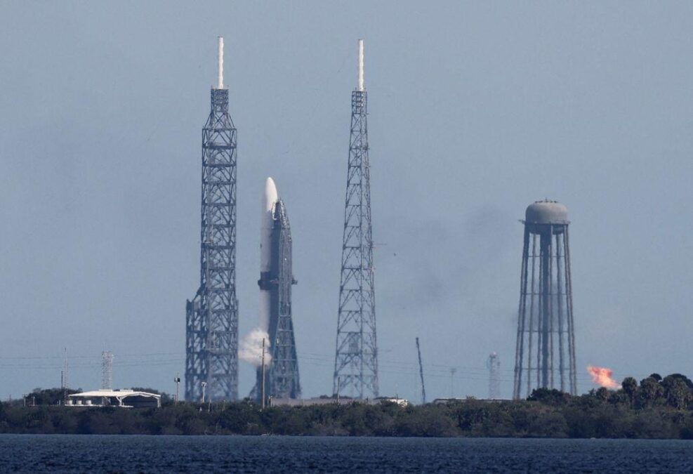 Launch pad with a rocket poised for takeoff, surrounded by launch towers and infrastructure, with smoke billowing from the base in preparation for lift-off.
